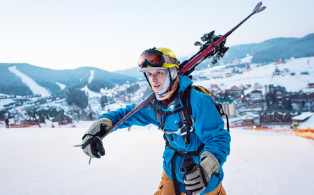 Close-up a guy skier in a blue jacket, a yellow helmet and glasses is holding sticks on his shoulder and staring into the distance against the backdrop of the ski resortの写真素材