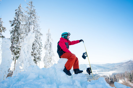 Man snowboarder sitting on top of a snowy hill with his snowboard enjoying stunning mountains view ski resort copyspace seasonal activity. Ski season and winter sports conceptの写真素材