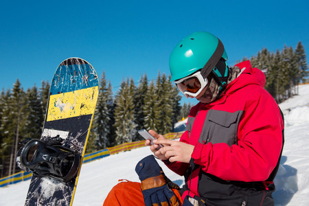 Close-up portrait of male snowboarder sitting in the snow, using his smart phone while resting in the mountains technology communication carrier mobility recreation sport lifestyleの写真素材