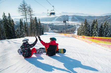 Snowboarders sit on the top of the ski slope under the ski lift let's high five to each other with a beautiful scenery of mountains and forests on a sunny morningの写真素材
