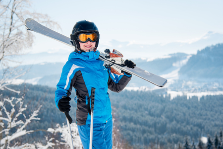 Portrait of happy female skier smiling to the camera, wearing skiing equipment, holding skis on the shoulder at winter ski resort copyspace happiness positivity vacation travellingの写真素材