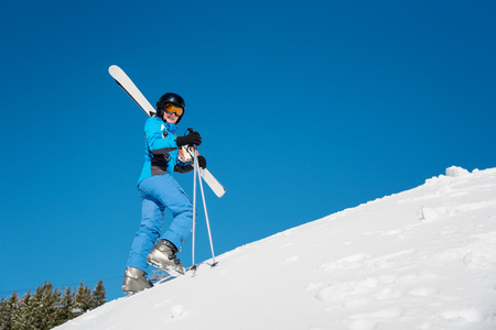 Low angle full length shot of a young female skier carrying her skis on a shoulder, looking to the camera while walking in the mountains. Blue sky, sunny winter day. copyspace freerideの写真素材
