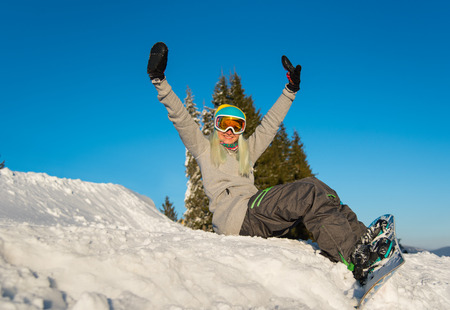 Low angle shot of a happy young woman snowboarder sitting on top of the slope, smiling to the camera, relaxing outdoors on the snow on a beautiful sunny winter dayの写真素材