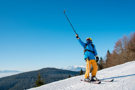 Full length shot of a professional skier taking a selfie using selfie stick posing on the slope. Blue sky, mountains and winter forest on the backgroundの写真素材