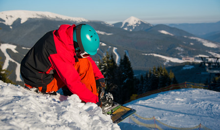 Male snowboarder sitting on the snowy slope, preparing for riding downhill on a beautiful sunny winter evening at ski resort Bukovel. Mountains, forests, ski slopes on the backgroundの写真素材