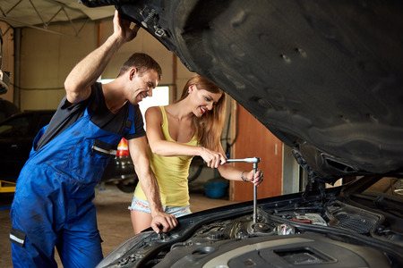 Young woman helps an auto mechanic repair a car in a garage. The girl unscrews the front right-hand stand. The guy holds the hood cover by handの写真素材