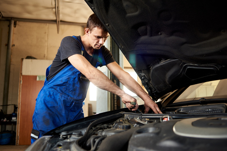 An auto mechanic in a dirty work uniform repairs a car in the garage. The bonnet is open, the guy looks carefully at the problemの写真素材