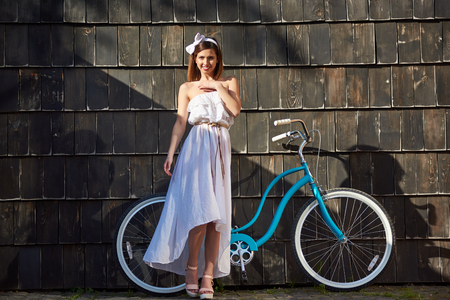 Lovely girl on a sunny day against a blue vintage bicycle and a dark wall. The girl is dressed in light clothes and looking at the camera with a smileの写真素材