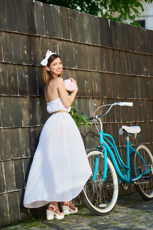 Happy young beautiful woman holding a bunch of peonies smiling to the camera posing elegantly near her bicycle outdoors romance romantic femininity sensuality.の写真素材