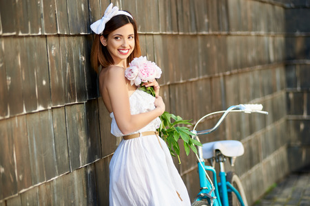 Happy woman holding peonies leaning against the wall and looking with a smile to the distance. In the background there is a retro bikeの写真素材