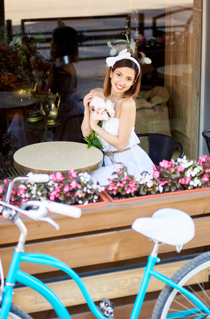 Attractive smiling female dressed in white sitting in a cosy cafe holding flowers and looking to the camera next her blue vintage bike. Pretty girl summer day happy life peonies coffeeshop exterior.の写真素材