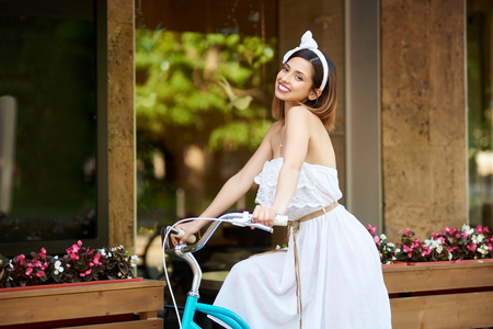 Smiling attractive female in white dress and bow headband riding a blue bike passing cafe decorated with flowers and wooden panels. Pretty girl happy enjoying sunny summer day cafe exterior.の写真素材