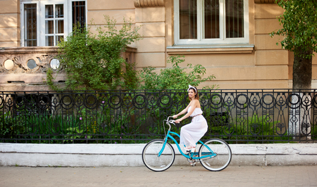 Lovely woman on a vintage bicycle is riding along the city street looking at the camera. The girl is dressed in a light dress and an accessory on her headの写真素材