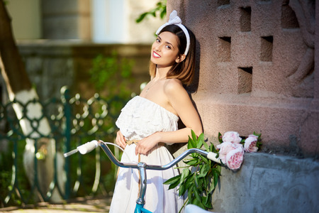 Attractive brunette in white dress leans on red old building wall with her blue vintage bike and peonies near. Beautiful female woman thinking flowers green photoset photoshoot.の写真素材