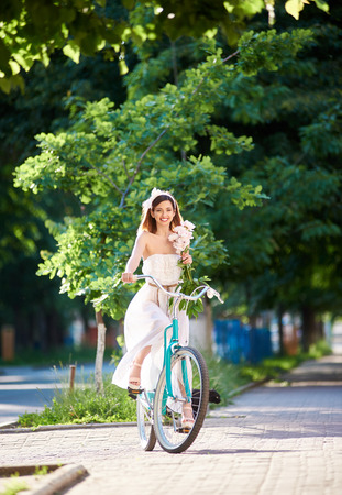 Vertical portrait of a beautiful young woman in white dress smiling cheerfully enjoying cycling in the park in summer happiness harmony positivity living leisure countryside.の写真素材