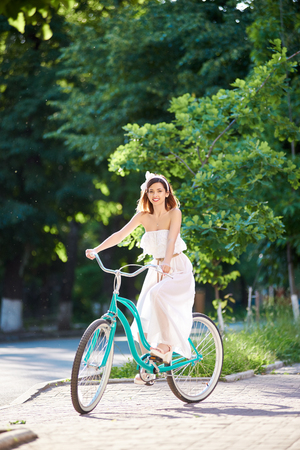 Beautiful woman in long white dress and bow headband smiling to the camera riding vintage blue bike in park on sunny summer day. Pretty female turquoise bicycle green trees happy smiling photoshoot.の写真素材