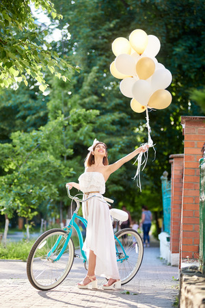 Happy young woman near vintage bicycle over city background and holding air balloons of tender shades at summer day.の写真素材