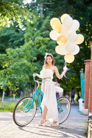 Pretty young woman in long white dress posing with colorful balloons in her hand near vintage blue bicycle on park alley on sunny summer day. Beautiful female turquoise bike smiling happy lifestyle.の写真素材