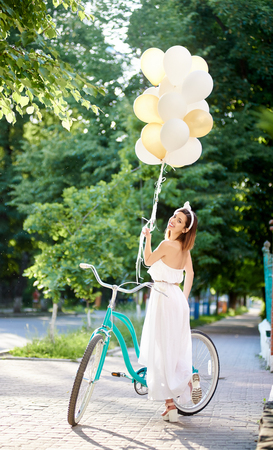 Rearview shot of a happy young beautiful woman smiling to the camera over her shoulder posing with a bicycle in the park holding colored baloons emotions youth active trip tourism happiness.の写真素材