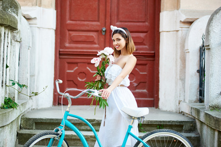Beautiful female in white dress posing with peonies near blue vintage bike in front of old beautiful porch with red antique doors. Pretty woman flowers historical building enjoying happy relaxの写真素材