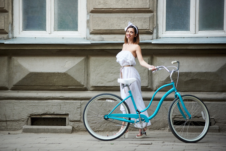 Pretty young female in long white dress posing with vintage blue bicycle with old historical building on the background. Summer day beautiful girl turquoise bike smiling happy enjoyingの写真素材