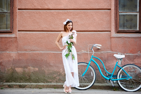 Pretty woman in long white dress and high heels posing with peonies and blue vintage bike with red historical building wall on background during summer day. Model flowers turquoise bicycleの写真素材