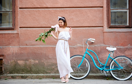 Young attractive elegant woman in a dress smelling bunch of peopnies standing on the street with her bicycle on the background copyspace lifestyle urban romantic weekend travelling.の写真素材