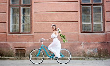 Smiling woman on a vintage bicycle is riding along the old city with peonies in her hand. The girl is dressed in a light dress and an accessory on her headの写真素材