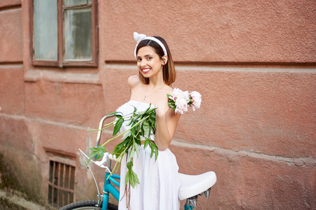 Horizontal portrait of a happy young woman smiling to the camera excitedly posing with a bouquet of peonies holding her bicycle copyspace city town village travelling emotions memories.の写真素材