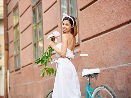 Close-up of a beautiful woman with peonies in hands in a light dress near a retro bicycle on the background of an old wall.の写真素材