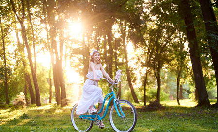 Beautiful girl in long white dress is holding peonies and riding blue bike in bright green park during sunny summer day. Smiling pretty female enjoying ride turquoise bicycle flowers treesの写真素材