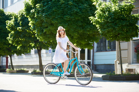 Smiling girl in white dress riding blue vintage bike and looking to the camera with green trees and flowerbeds in background during sunny summer day. Pretty female turquoise bicycle happy relax cityの写真素材