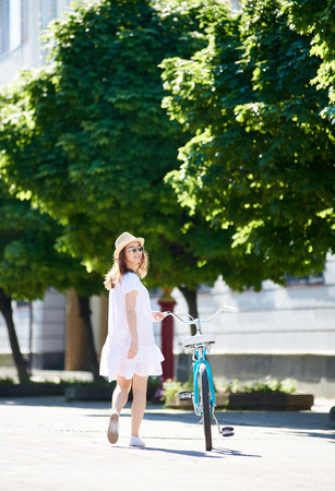 Lonely girl comes with a retro bicycle on the sunny city street with green spaces. The girl turned to look at the camera, she is dressed in a light dress, sunglasses and a hat on her headの写真素材