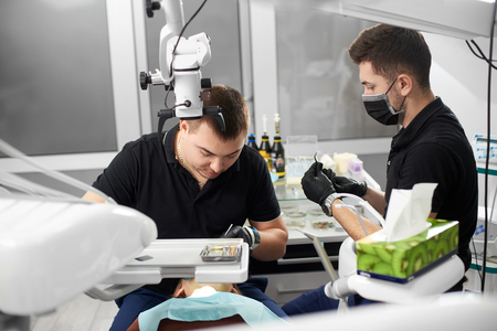 Two male dentists in black uniforms are working with dental instruments while curing teeth of a female patinet of a white dental clinicの写真素材