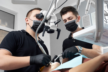 Male dentist looks in a microscope curing patient's teeth, another male doctor assists himの写真素材