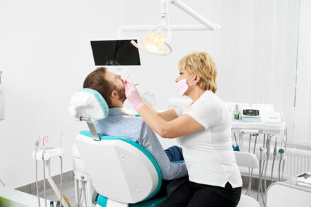 Female dentist in mask with mirror checking patient man teeth up at modern dental clinic officeの写真素材