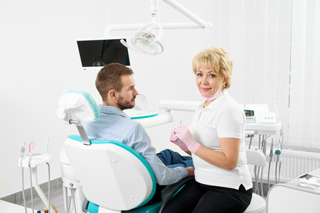 Beautiful female dentist is looking at the camera holding medical instruments and being ready to cure the teeth of young male client of the dentistry.の写真素材