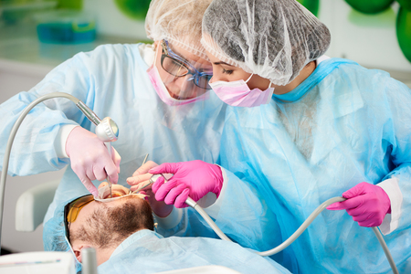 Two dentists in blue robes are scaling and polishing the teeth of a male client of a dental office.の写真素材