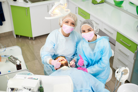 Photo from above doctor woman and her assistant dressed in a one-time uniform work with a male patient in a modern dentistry office and shows gesture classの写真素材