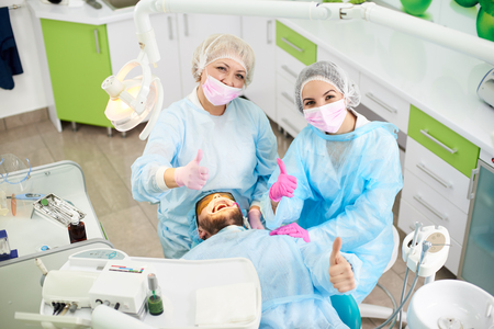 Happy and smiling dentists and their male client showing all thumbs up while successful dental treatment in a dental office.の写真素材