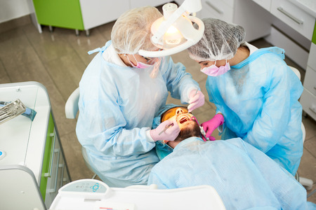 Doctor woman and her assistant dressed in a one-time uniform work with a male patient in a modern dentistry office. Photo from aboveの写真素材