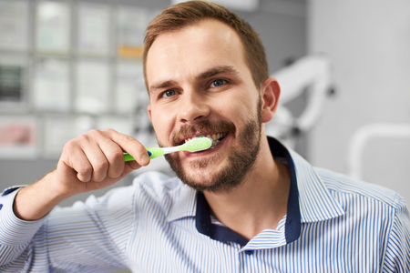 Young attractive male is smiling to the camera while cleaning his teeth with a tooth brush in a modern dental office.の写真素材