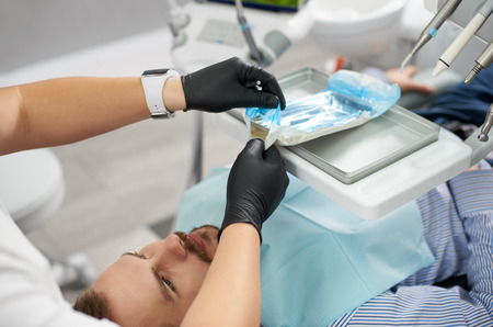 Professional in black rubber gloves is getting ready to attend the teeth of a young male client of a dental office.の写真素材