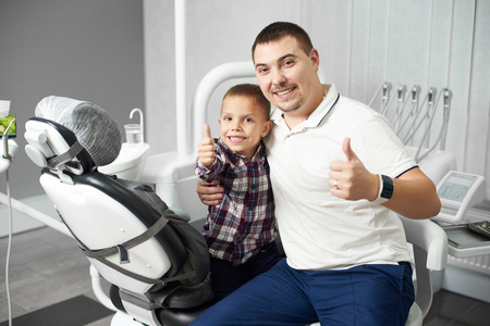 Male dentist and his young child client are sitting together hugging and showing thumbs up in a dental office, both being satisfied and happy after the dental treatment.の写真素材