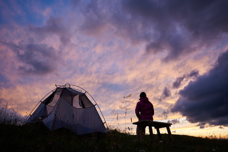 Hiker woman sitting on a bench near a tent and looking at incredible view of sunset evening sky with purple clouds. Young tourist girl alone in cold weather near the campsite outdoors. Travel concept.の写真素材