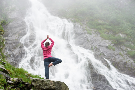 Pretty young girl standing in a yoga pose near big powerful waterfall. Beautiful woman meditating in wild serene nature. Traveling to the waterfall Balea in Romanian Fagarash mountains.の写真素材