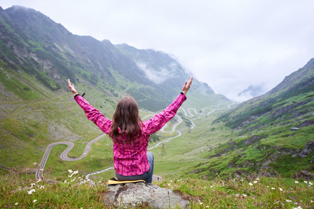 Woman tourist sits with her arms wide open and looks up against background of Transfagarashan Highway, the most beautiful road in Europe, Romania. Back viewの写真素材