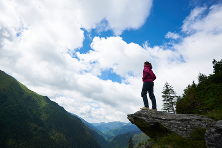 Upward shot of young female tourist standing on top of the rock admiring blue sky and amazing scenery with green grassy slopes and mountains in Romania. Woman climber travel trees beautiful viewの写真素材
