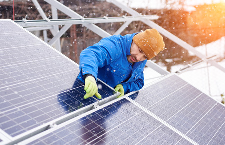 Guy with the help of tools installs solar panels in snowy weather. He is dressed in uniform and...