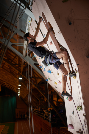 Reaching the top. Attractive sportsman putting efforts to boulder artificial climbing wall in bouldering gym indoors. Motivation strong healthy male man climber exercise hobby activity leisure timeの写真素材